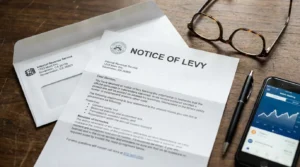 An IRS notice of levy letter on a desk next to reading glasses and a phone showing a banking app, representing what a notice of levy means for taxpayers in 2026